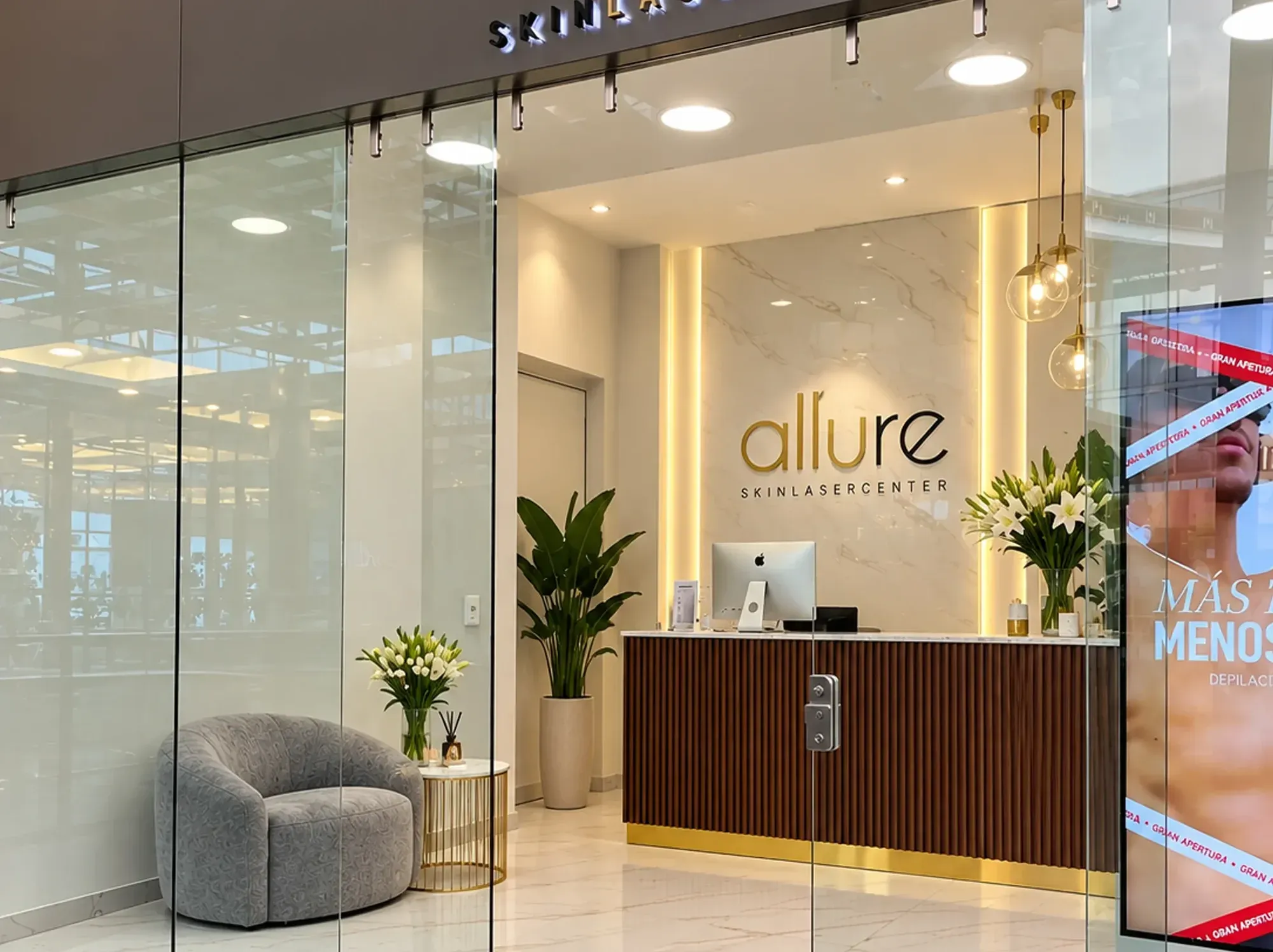 Recepción de Allure Skin vista desde la entrada: logo dorado sobre pared de mármol, mostrador de madera, sillón gris y flores blancas