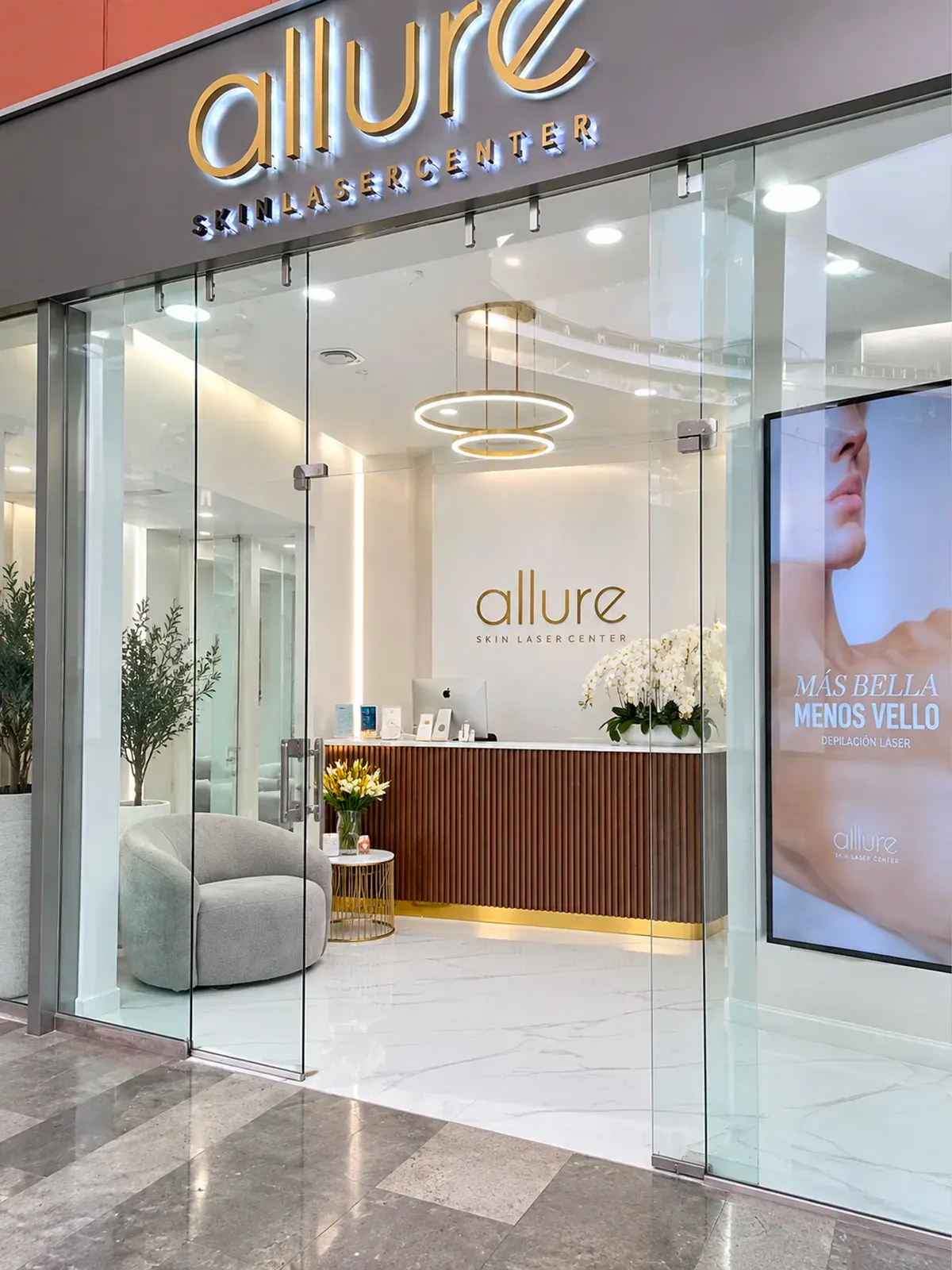 Fachada de Allure Skin en Plaza Punto Sur, Guadalajara: letrero dorado iluminado, puertas de cristal, recepción con mostrador de madera y orquídeas blancas