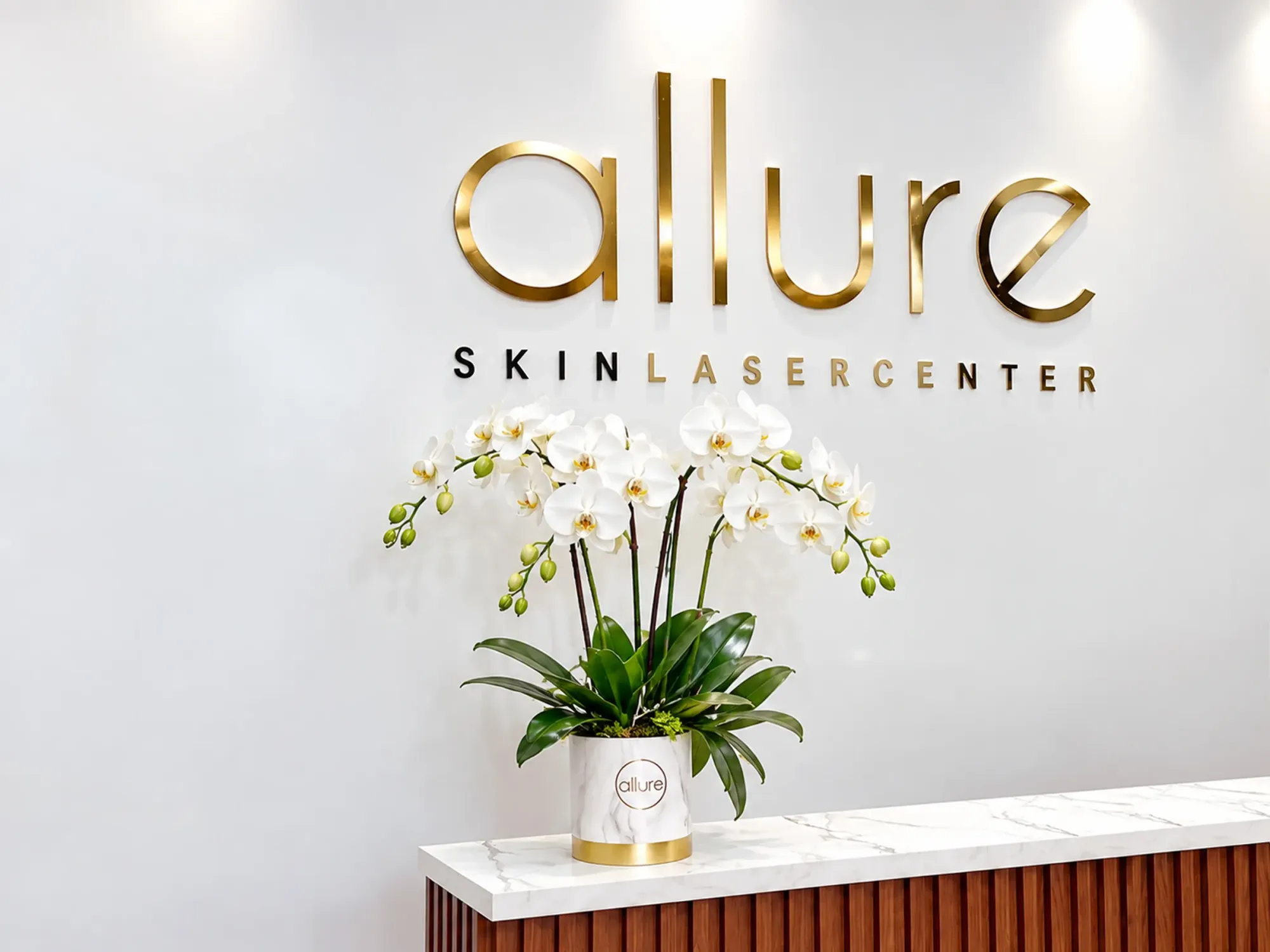 Pared de recepción de Allure Skin con el logo dorado y un arreglo de orquídeas blancas sobre el mostrador de mármol